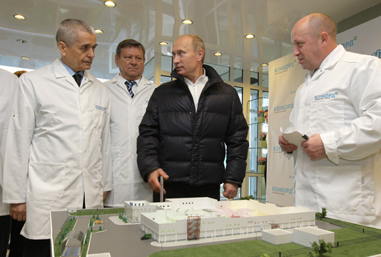 Then Prime Minister Vladimir Putin with factory director Yevgeny Prigozhin, looked over the factory's production chain and was shown new vending machines that accept cards (government.ru, CC BY 4.0 <https://creativecommons.org/licenses/by/4.0>, via Wikimedia Commons). Source: https://commons.wikimedia.org/wiki/File:Vladimir_Putin_tours_Yevgeny_Prigozhin%27s_Concord_food_catering_factory_02.jpg