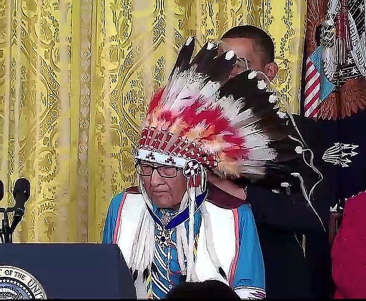 Joseph Medicine Crow-High Bird at the 2009 Medal of Freedom ceremony, the award is seen around his neck, and President Obama is behind him, wrapping the award around his neck. (<a href="https://commons.wikimedia.org/wiki/File:Joseph_Medicine_Crow-High_Bird_-_Aug_12_2009_Presidential_Medal_of_Freedom_-_with_Obama_and_award.jpg">US Government</a>, Public domain, via Wikimedia Commons)