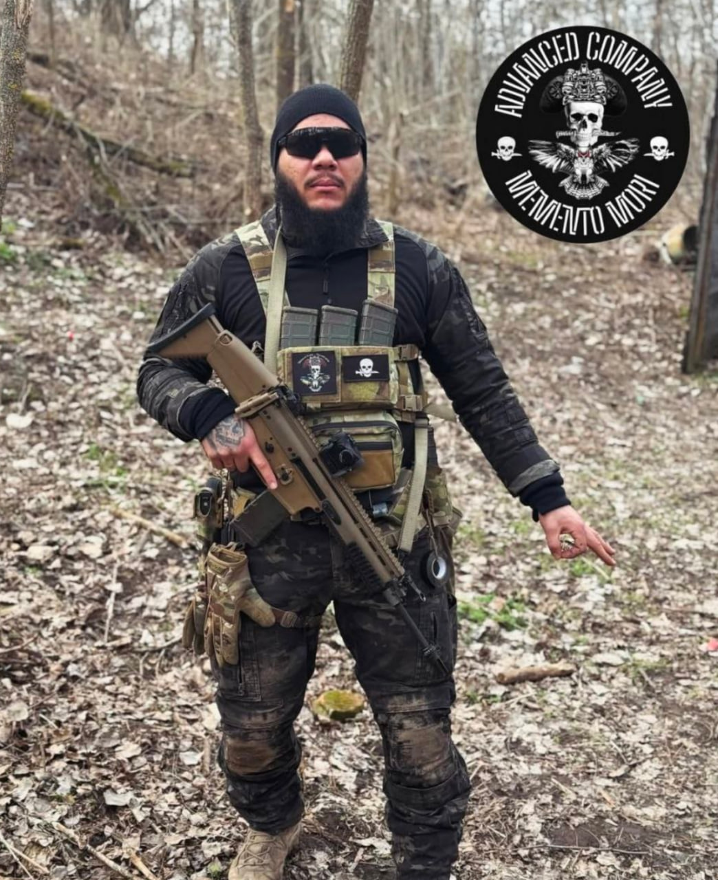 A man wearing tactical gear and sunglasses stands in a wooded area holding a rifle, with a circular “Advanced Company” logo visible in the image.