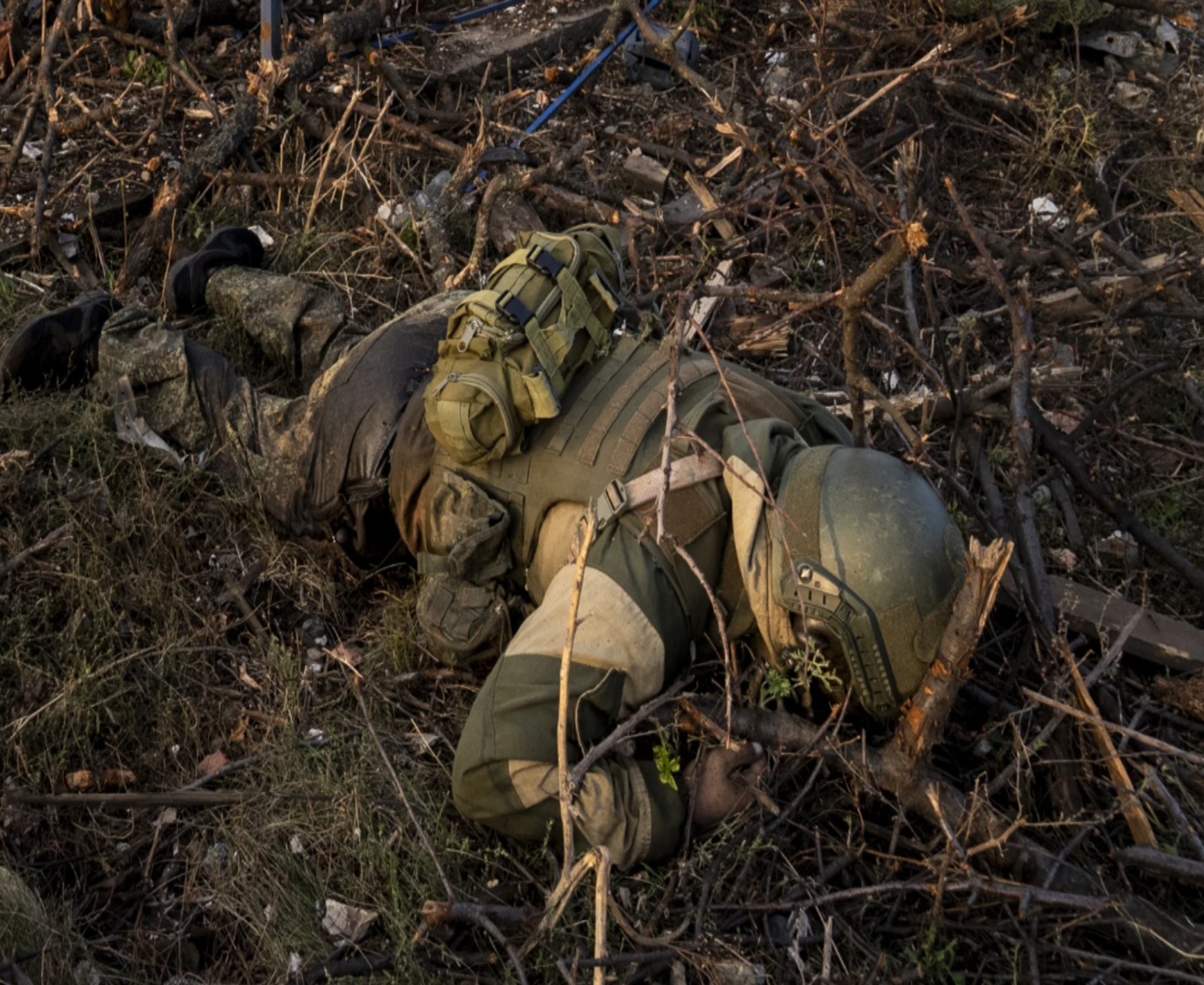 A Russian soldier wearing a helmet and tactical gear lies prone on the ground among branches and debris during training outside Bakhmut.