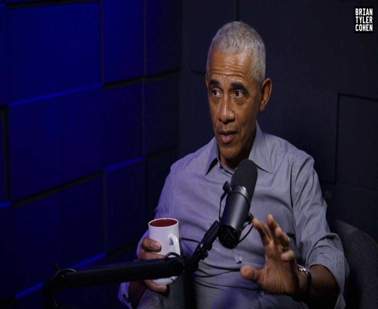 Former President Barack Obama sits in a podcast studio holding a coffee mug while speaking into a microphone during an interview with Brian Tyler Cohen
