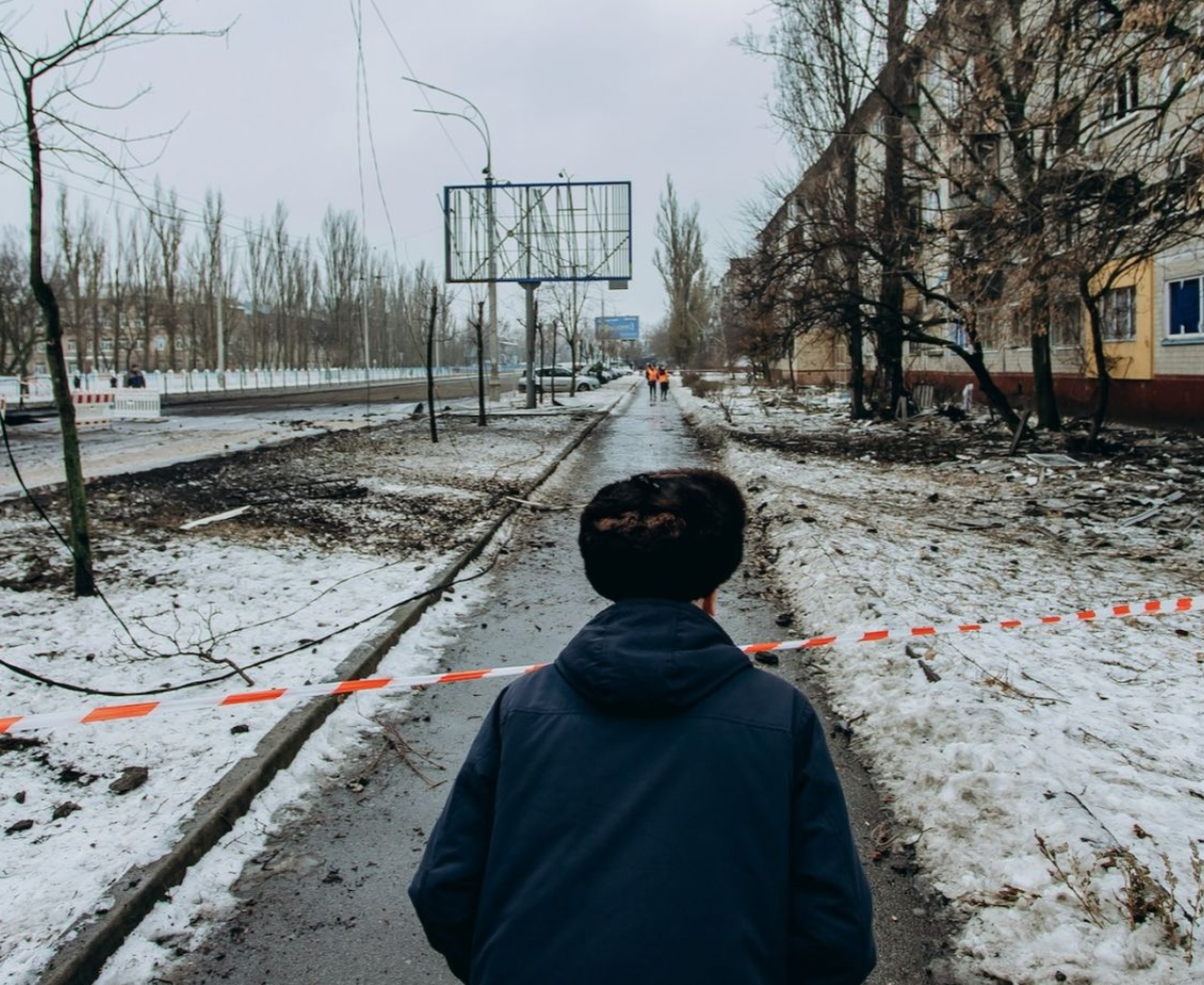 Civilian walking along a damaged, snow-covered street in Ukraine with debris and destroyed buildings after Russian strikes on energy infrastructure.