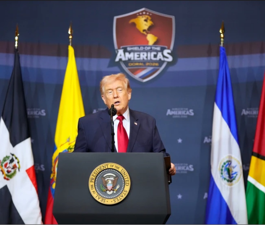 President Donald Trump speaks at a podium during the Shield of the Americas Summit in Doral, Florida, with Latin American flags behind him.